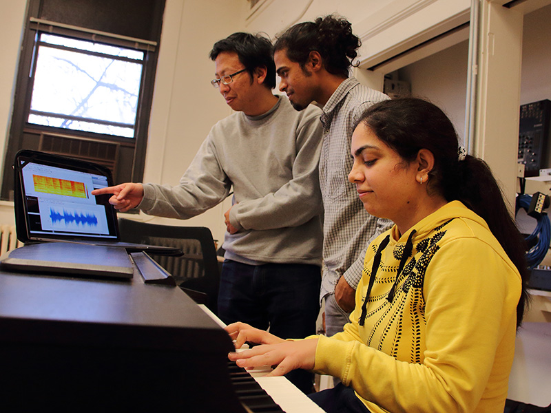A person plays at a keyboard while two others look at a computer.
